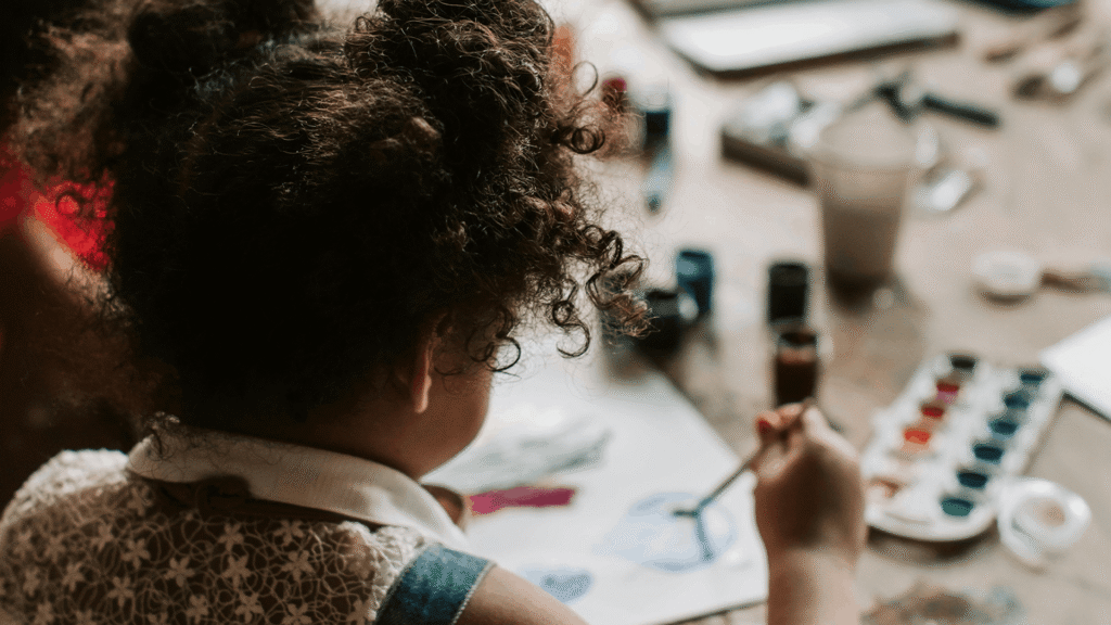 Over-the-shoulder view of a child painting with watercolors at a table.