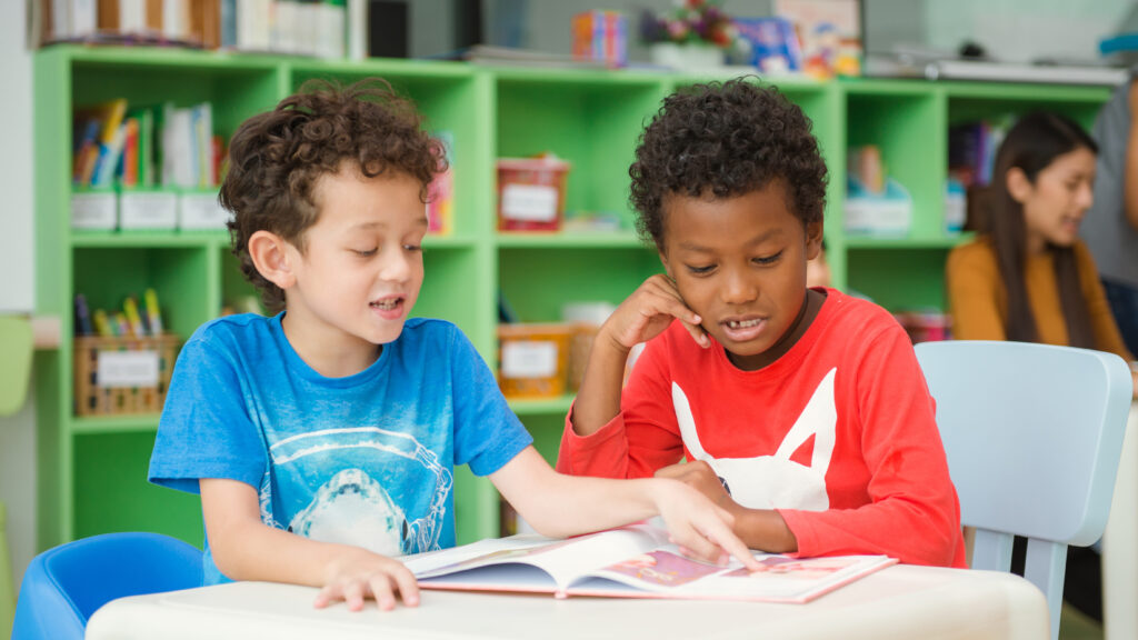 Two children sitting at a table in a library reading a book. One child points at a picture in the book, both children look engaged with what they are reading.