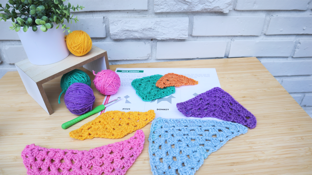 A desk. On the desk are tangram shapes crocheted in different colors and a Storytelling with Shapes puzzle page. On the back-left side of the desk is a plant in a white pot on top of a light wooden stand. Next to and below the plant are colorful balls of yarn and a crochet hook The background is a white brick wall.