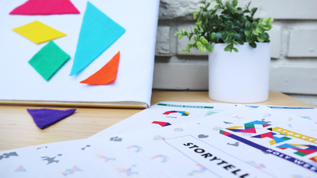 A close up of Storytelling with Shapes  adapted for use on a felt board. Colorful tangram shapes hang on the board and one lays on the table. In the background there is a plant in a white pot and a white brick wall.