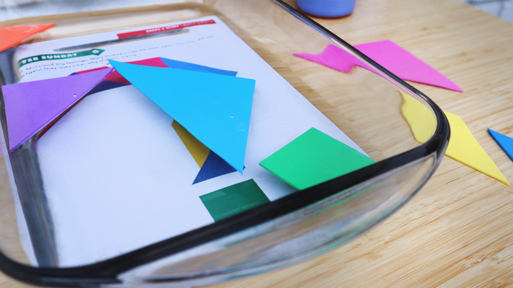 A close up on a glass baking pan filled with an inch of water. Under the pan is a Storytelling with Shapes puzzle page, floating on the water are colorful tangram shapes cut from foam sheets. 