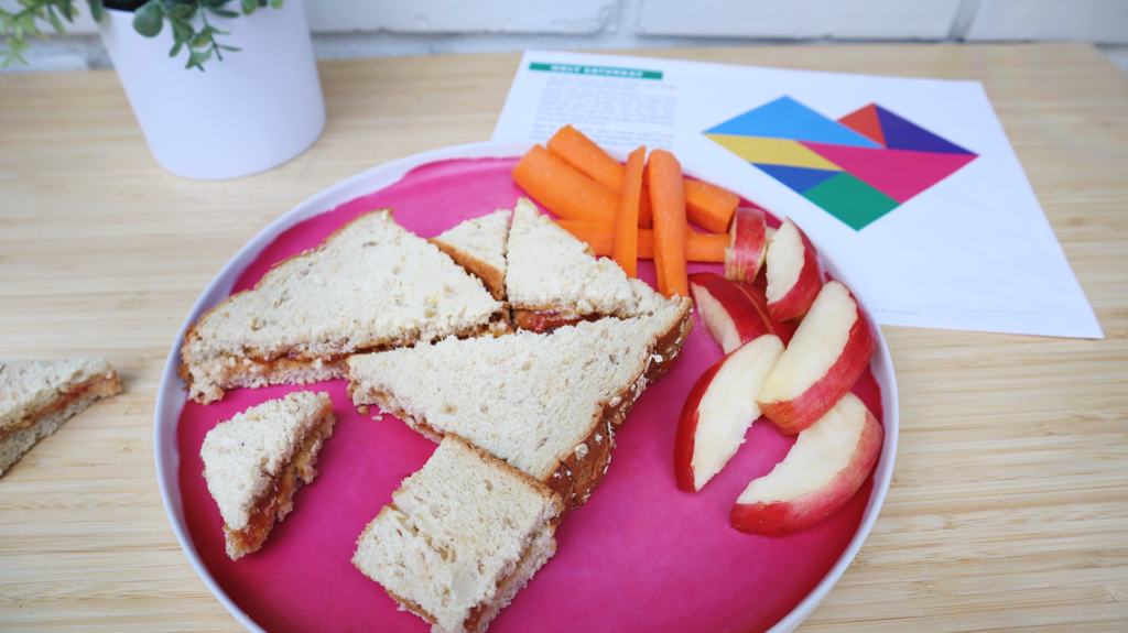 A close up on a circular pink plate placed on a desk. On the plate is apple slices, carrot sticks, and a peanut butter and jelly sandwich cut into tangram shapes. Under the plate is a Storytelling with Shapes puzzle page and at the back-left of the desk is a plant in a white flower pot.