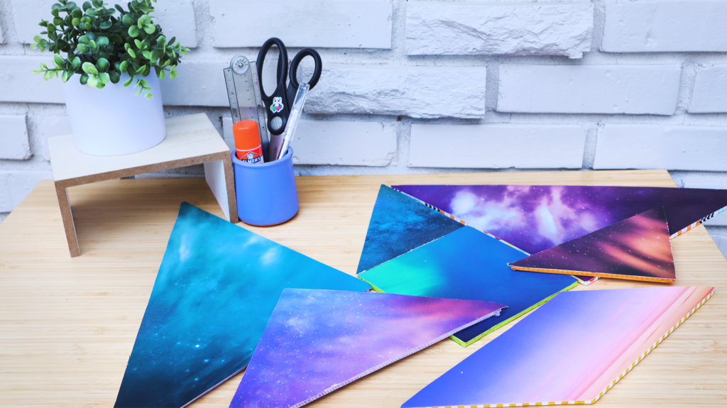 A desk. On the desk are tangram shapes cut out of a large foam board, each shape is covered by a colorful image of outer space. On the back-left side of the desk is a plant in a white pot on top of a light wooden stand and a small, periwinkle clay pot holding a ruler, scissors, glue stick, and craft knife. The background is a white brick wall.