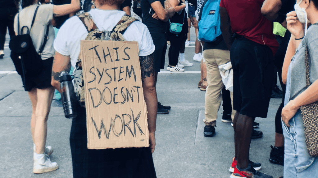 Person at a protest wearing a sign that says “This system doesn’t work.”