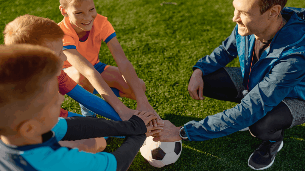 Male coach crouching on a soccer field with a group of boys, all placing their hands in the center for a team cheer