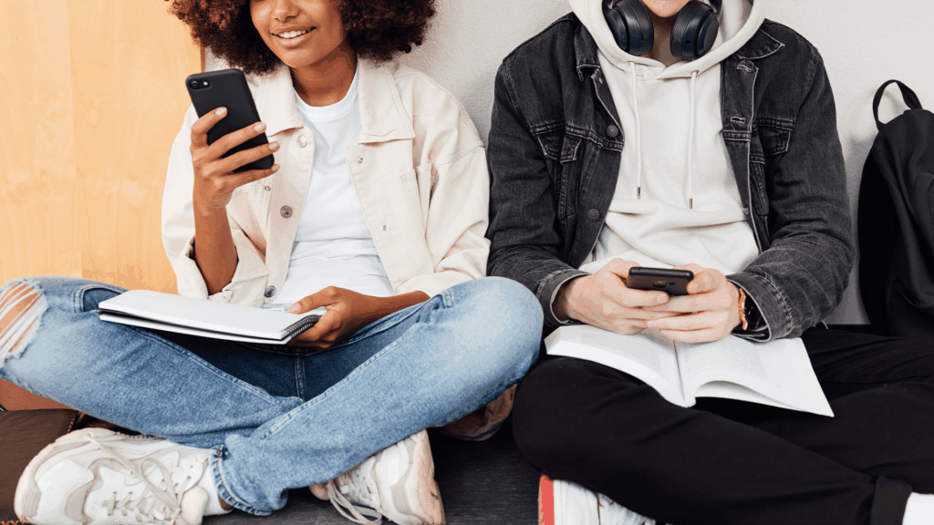 Two teenagers sitting on the floor, smiling and looking at their smartphones while holding open notebooks, symbolizing how teens balance schoolwork and social media use.