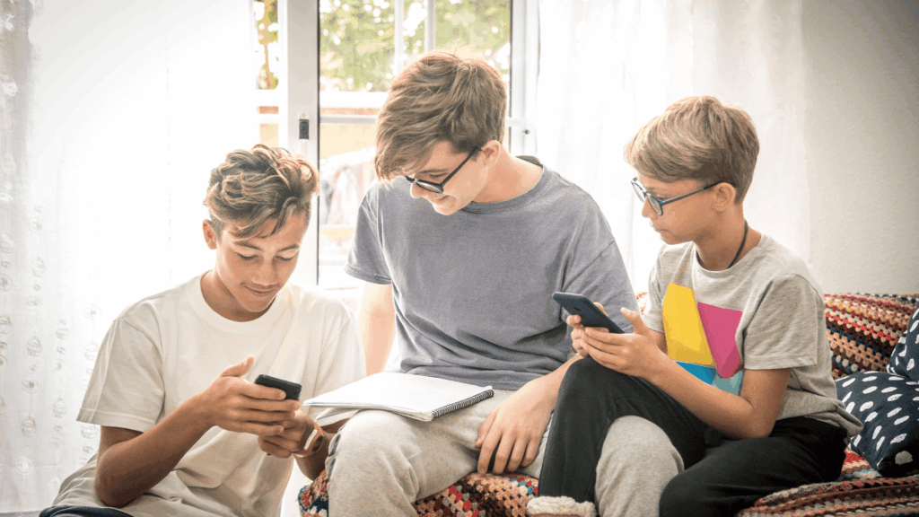 Three teenage boys sitting on a couch together, smiling and looking at smartphones and a notebook

