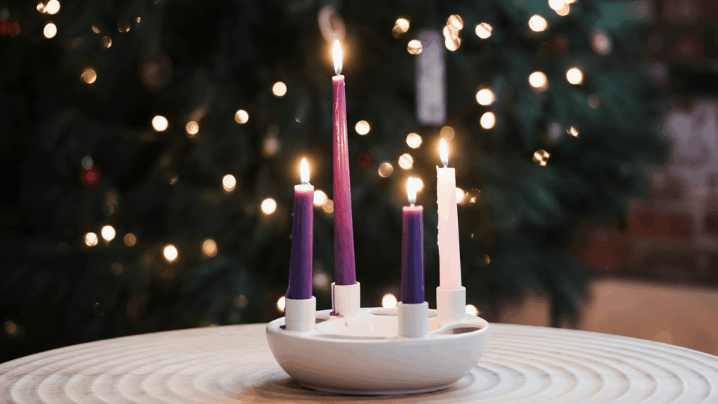 Four lit candles—three purple and one pink—sit in a white Advent wreath holder on a table, glowing softly against a blurred Christmas tree background with twinkling lights.