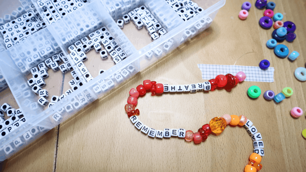 Red, orange, and yellow beads spelling Remember and Loved being strung together for a Beaded Examen craft, surrounded by alphabet beads and colorful supplies.