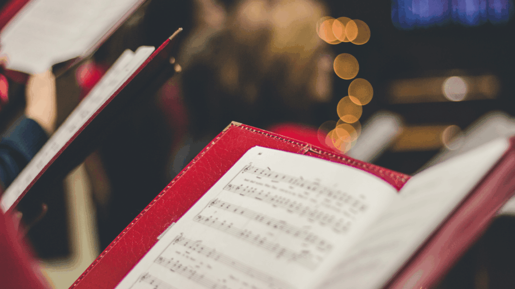 Close-up of an open hymnal with sheet music, held by a choir member during worship, with warm candlelight and blurred congregation in the background.