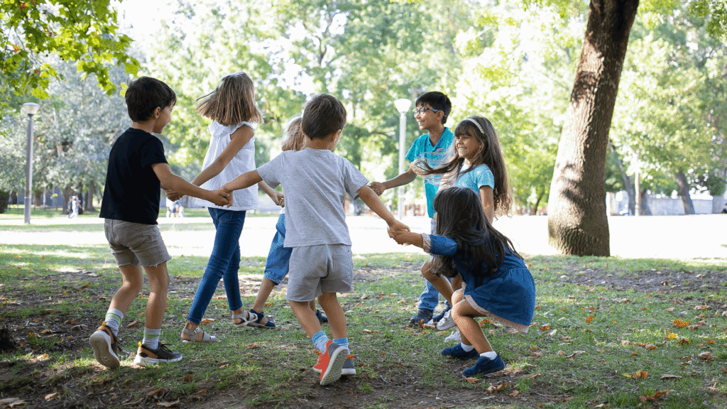 A diverse group of young children hold hands and play together in a circle in a park.