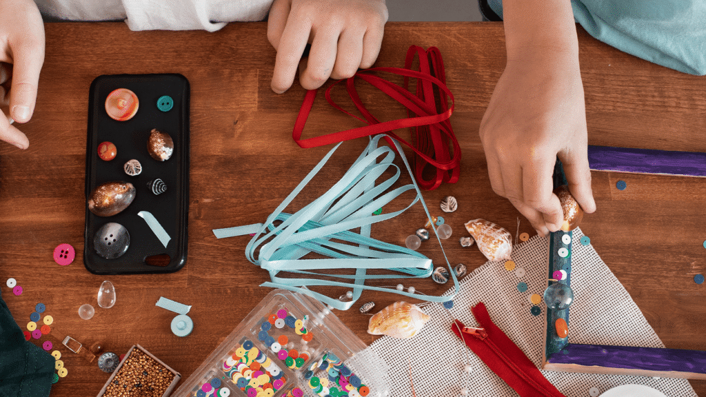 Children’s hands arranging ribbons, shells, and colorful craft supplies on a wooden table during a creative activity.