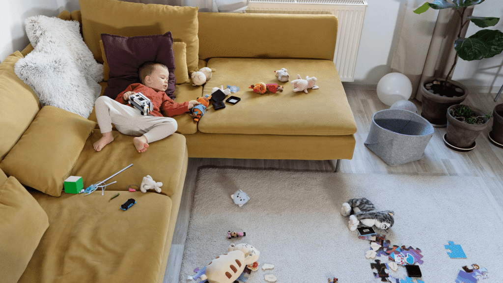 Child asleep on a couch surrounded by toys after a day of play, capturing the messy reality of parenting.