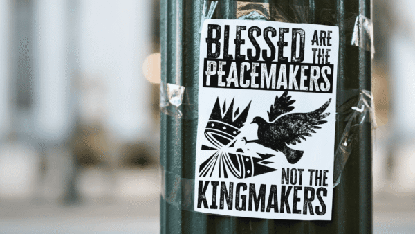 Protest sign taped on a green pillar against a soft blur urban background. The sign reads "blessed are the peacemakers not the king makers" in distressed font with the image of a dove breaking a crown