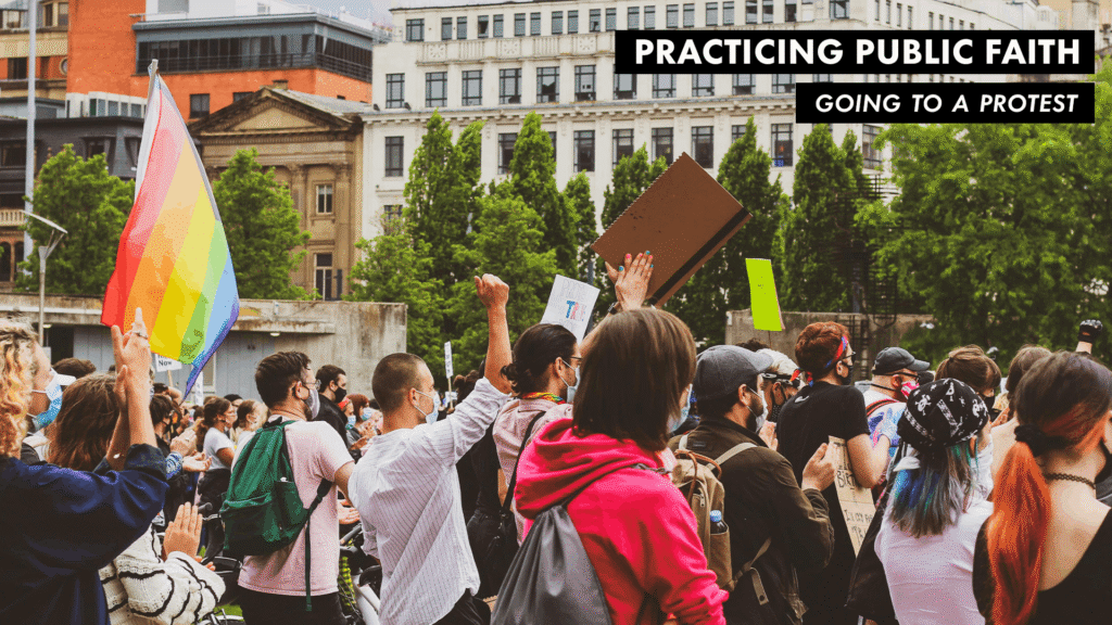 Crowd of people gathered at a peaceful protest holding signs and a rainbow flag in a city park.