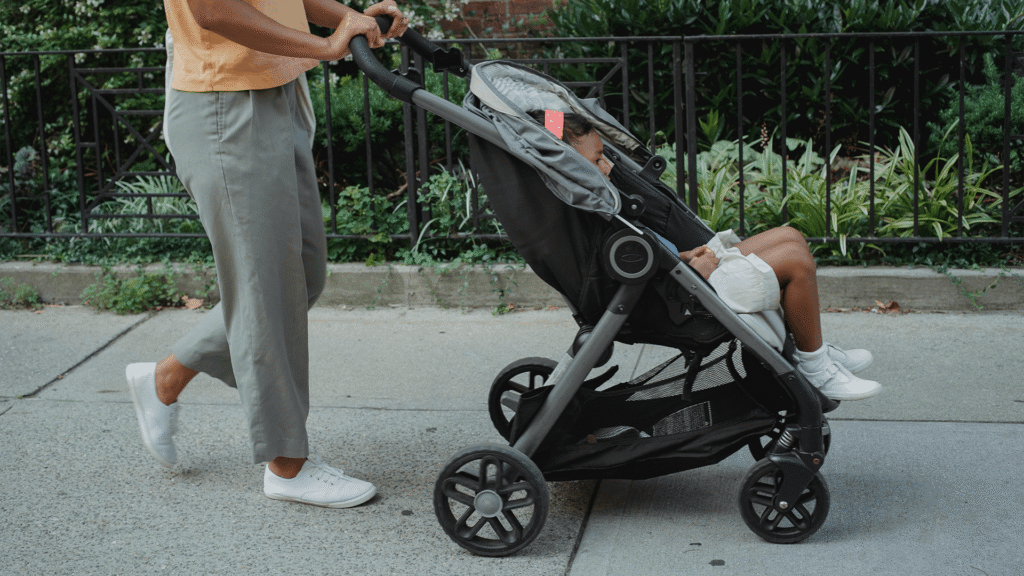 Caregiver pushing a stroller with a child along a neighborhood sidewalk on a sunny day.