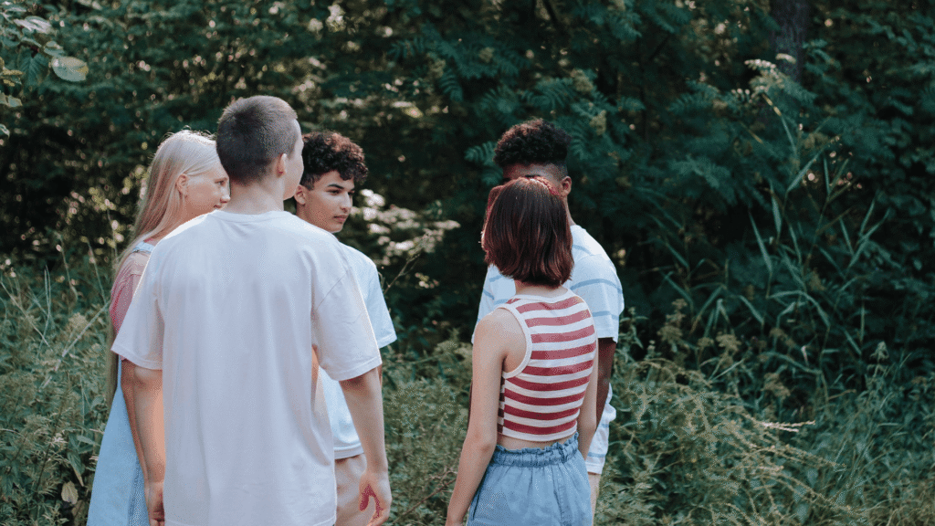 A small group of teenagers stand together outdoors in conversation, surrounded by trees, listening and talking with one another.