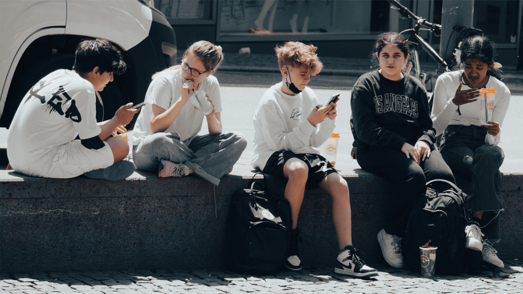 A group of teenagers sit together outdoors, with several focused on their smartphones while others look around, illustrating how phones can divide attention even when people are together.