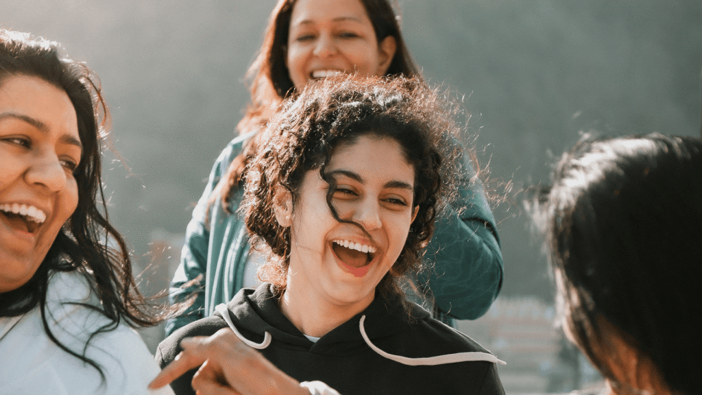 A diverse group of teenagers laugh together outdoors while talking in a circle, enjoying face-to-face conversation without phones.