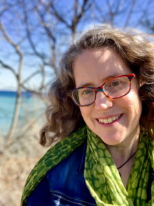 Author Laura Alary smiles outdoors beside a lake on a sunny day, wearing colorful glasses, a green patterned scarf, and a denim jacket.