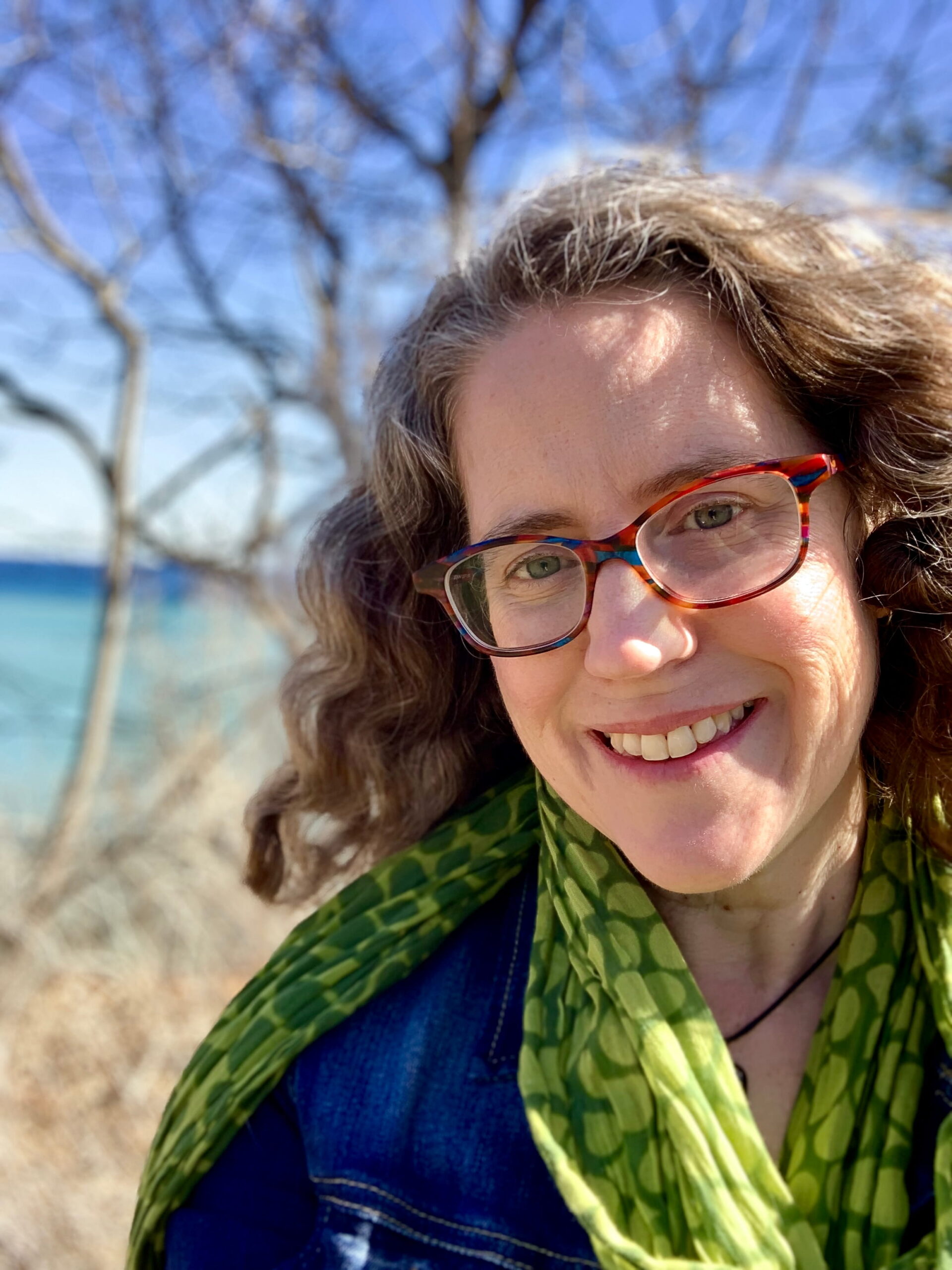 Author Laura Alary smiles outdoors beside a lake on a sunny day, wearing colorful glasses, a green patterned scarf, and a denim jacket.