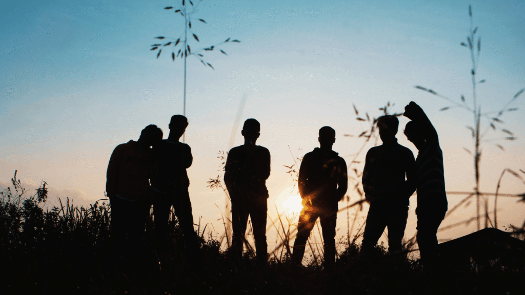 Silhouettes of several teenagers stand together in a grassy field at sunset, with the sun shining between them.