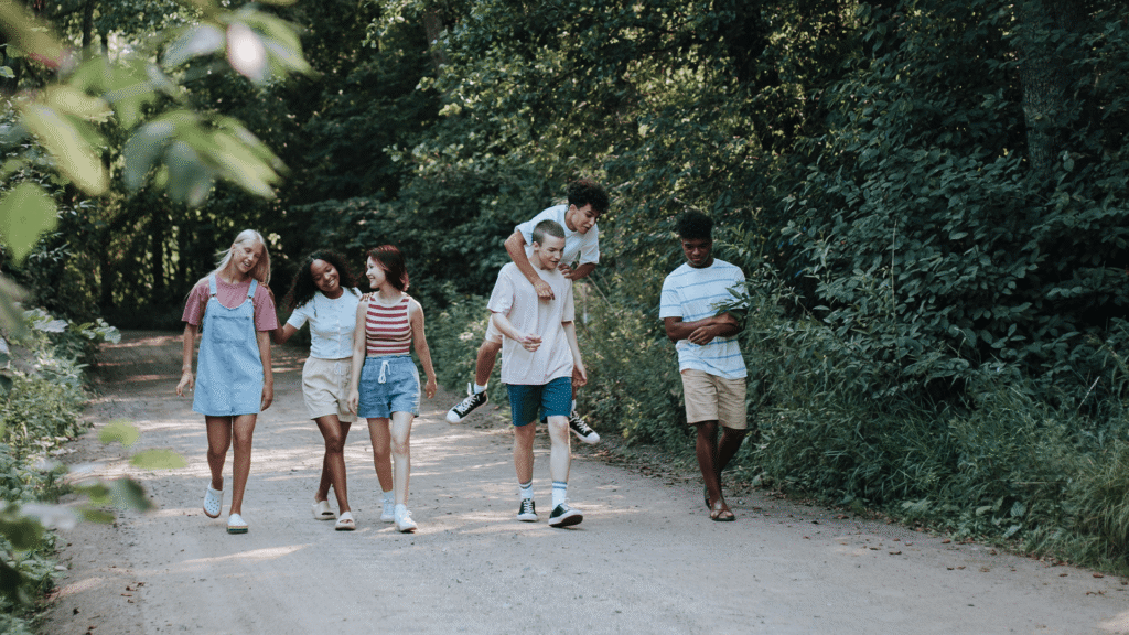 A diverse group of teenagers walk together along a wooded path, laughing, talking, and enjoying time outdoors.