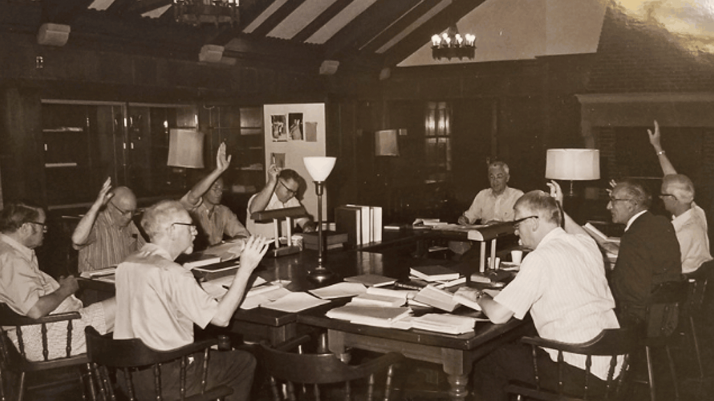 Black-and-white archival photograph of men gathered around a large table covered with books and papers, several raising their hands.